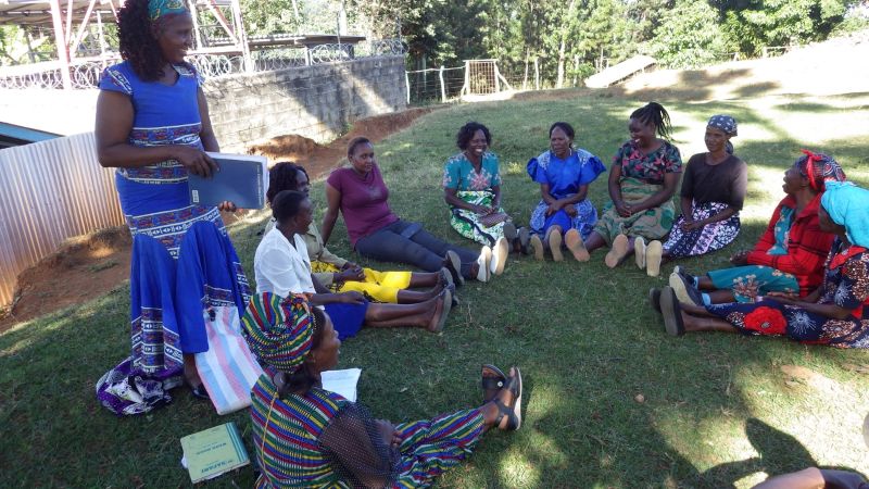 屋外で円になって座る女性たちと、その横に立つ女性の姿